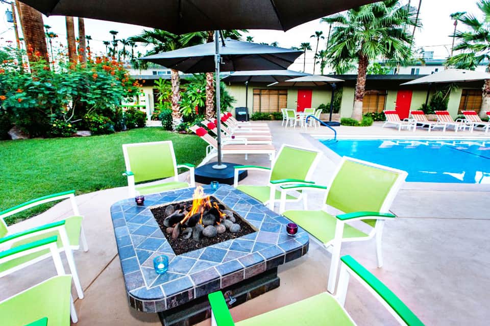 Resort pool area with lime green lounge chairs, blue umbrellas, tile fire pit, palm trees, and red-door cabanas
