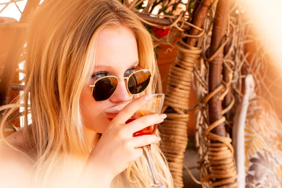 Woman wearing sunglasses enjoying a tropical drink with palm trees and woven details in background