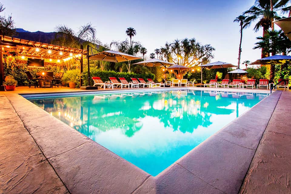 Resort pool at dusk with illuminated pergolas, lounge chairs, and palm trees reflecting in turquoise water