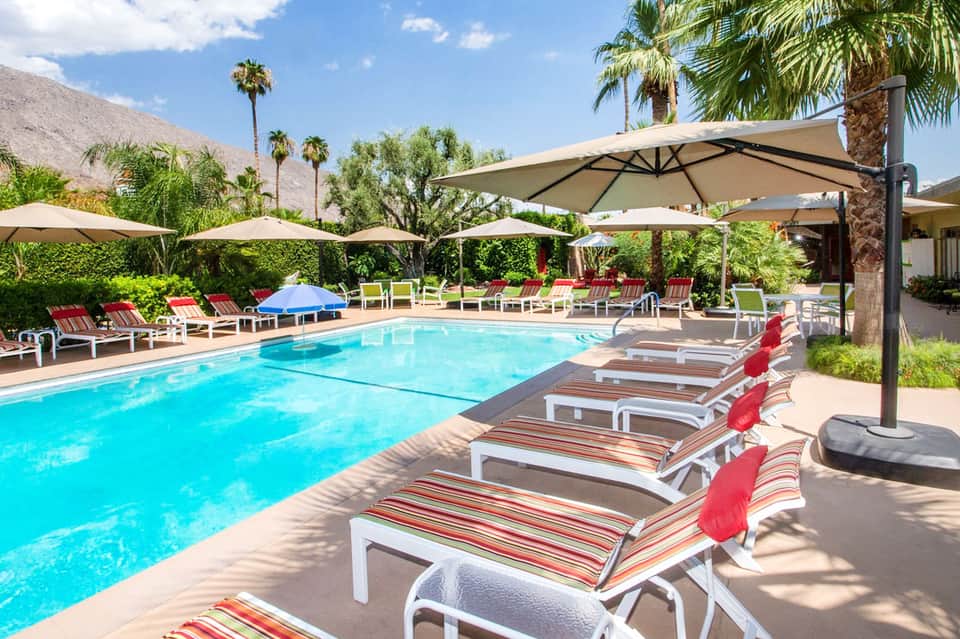 Resort pool with turquoise water, striped lounge chairs, large umbrellas, and palm trees with desert mountains in background