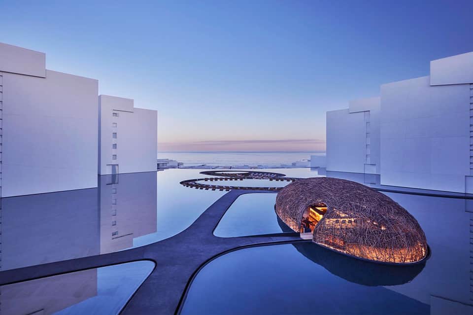 Luxury rooftop infinity pool at dusk with woven dome structure and city lights reflecting in water