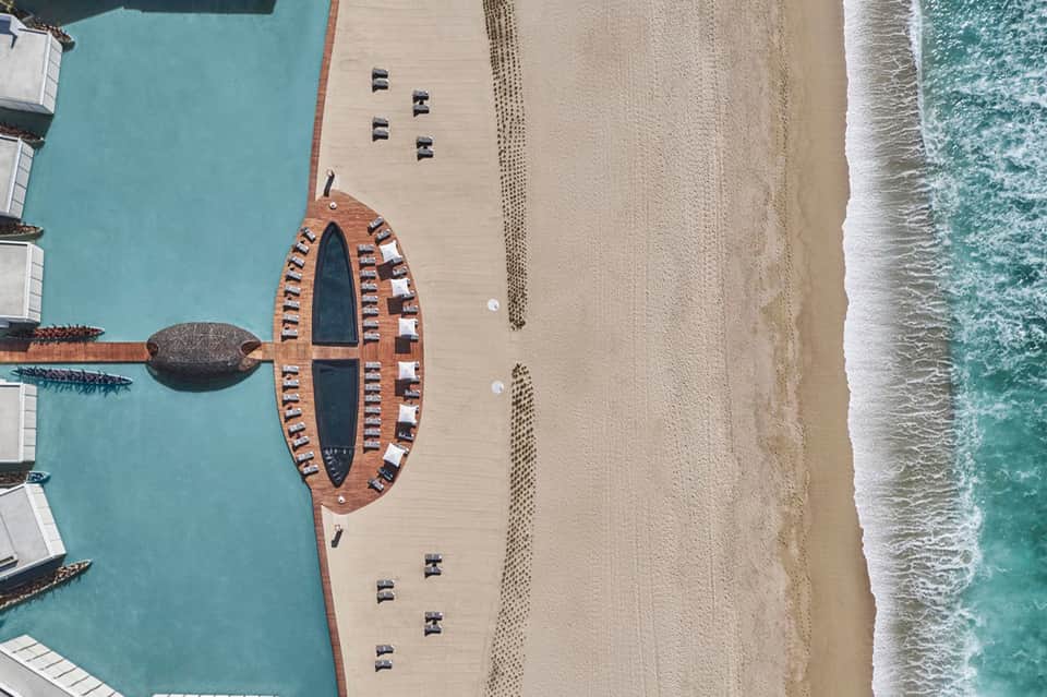 Aerial view of beachfront resort with oval-shaped pool, wooden dock, and turquoise water