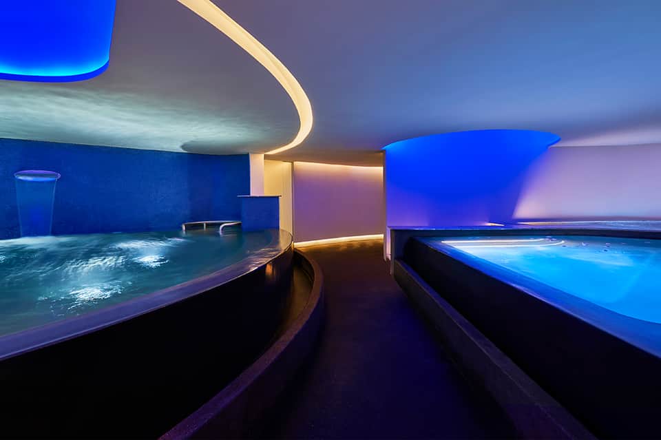 Modern spa with blue-lit hot tubs, curved architectural ceiling, and ambient warm lighting