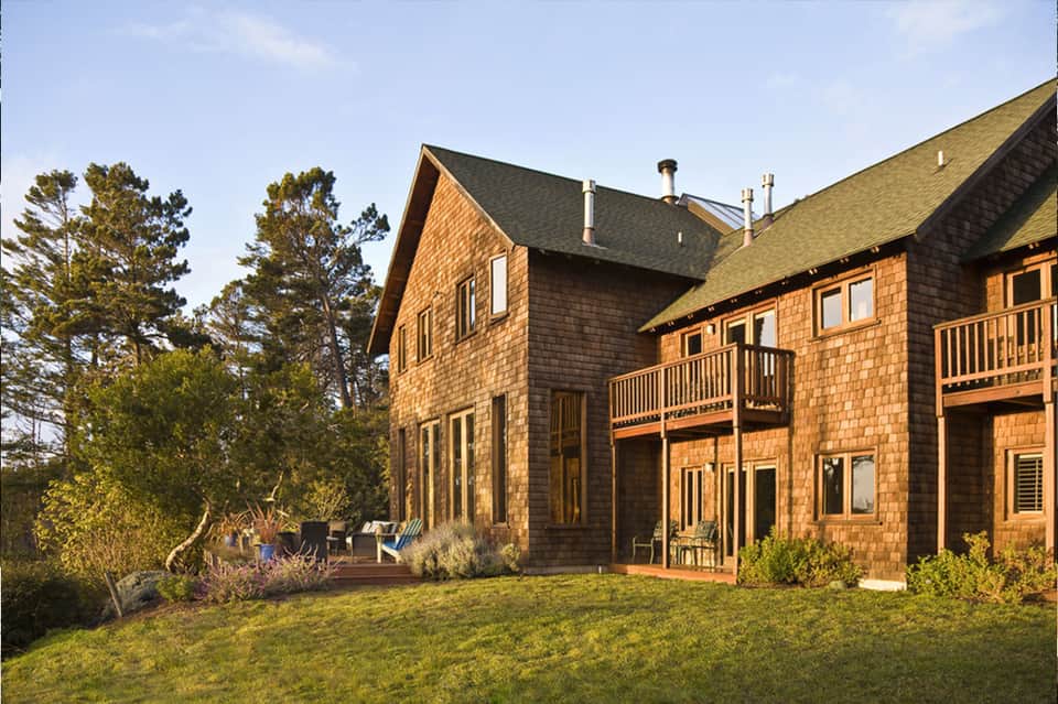 Modern brick residential home exterior with multiple decks and manicured lawn surrounded by pine trees