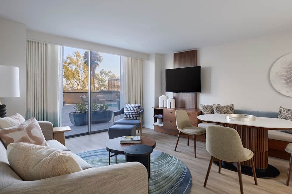 Contemporary hotel suite with neutral furnishings, flat-screen TV, balcony, and city views through large windows