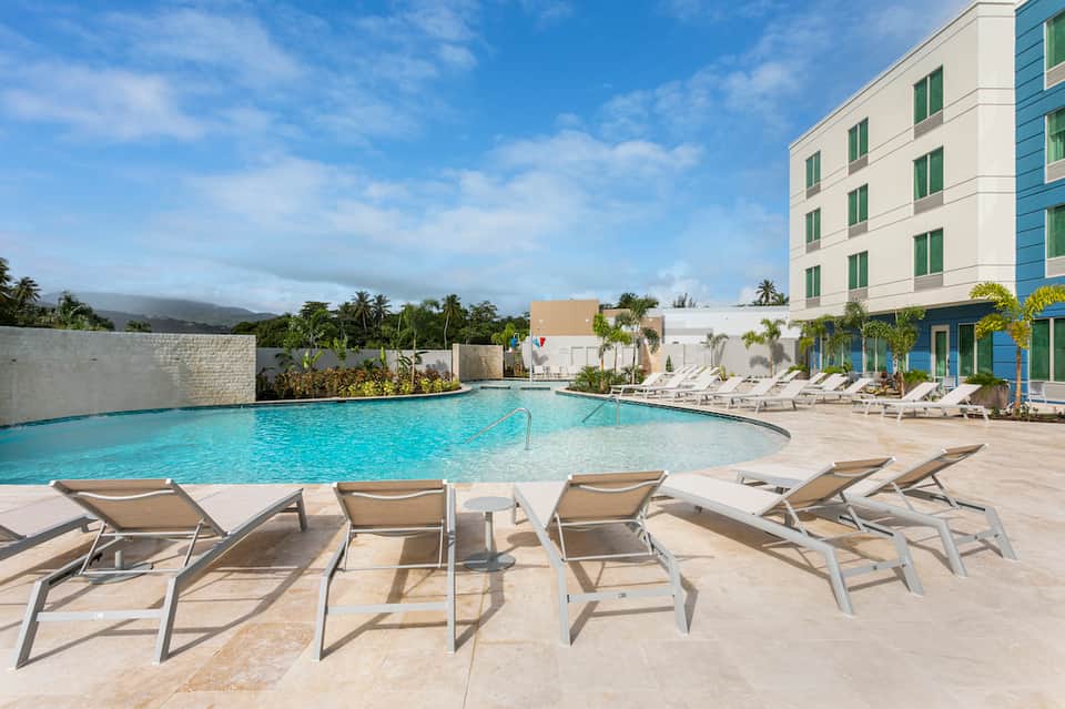 Resort pool area with turquoise water, tan lounge chairs, hotel building, and mountain backdrop