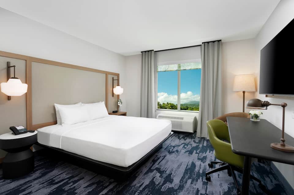 Modern hotel bedroom with queen bed, desk area, and large window overlooking mountains and blue sky
