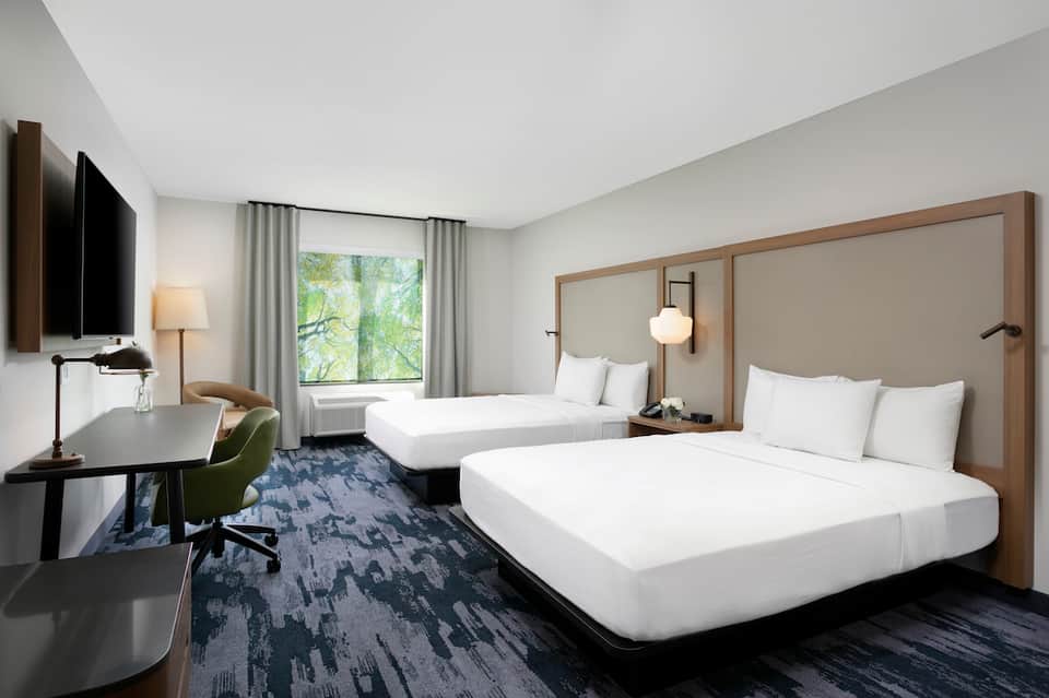 Modern hotel room with two queen beds, wood headboards, desk area, and window with natural light