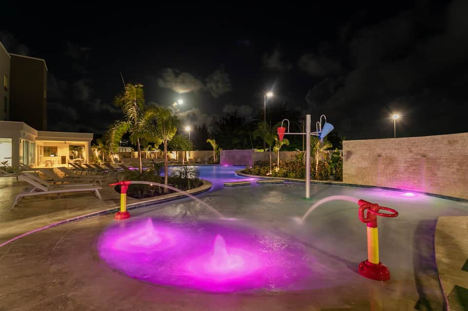 Resort spa area at night with illuminated magenta hot tub, palm trees, lounge chairs, and ambient lighting