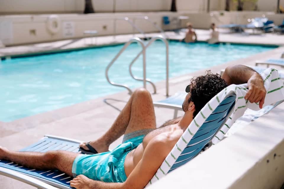 Man relaxing on lounge chair by indoor swimming pool with metal railings and white deck