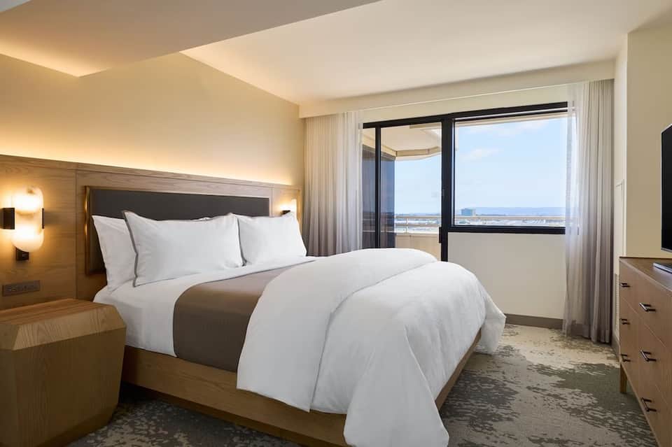 Modern hotel bedroom with queen bed, wood headboard, large windows, and scenic water view