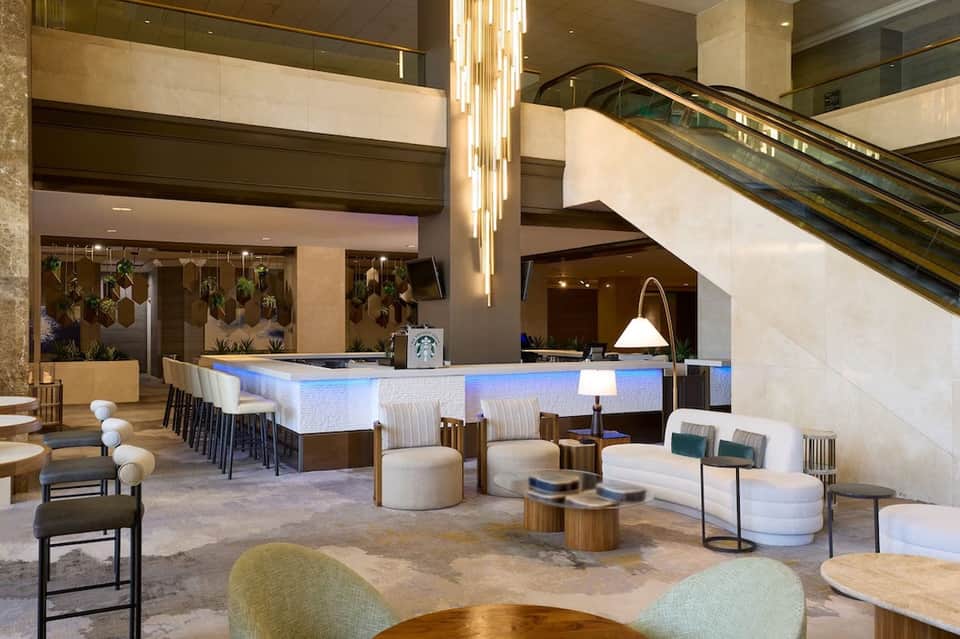 Modern hotel lobby with multi-level design, blue-lit bar, hanging plants, and contemporary furniture