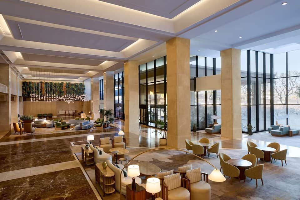 Luxury hotel lobby with tall columns, modern lighting, seating areas, and floor-to-ceiling windows overlooking water