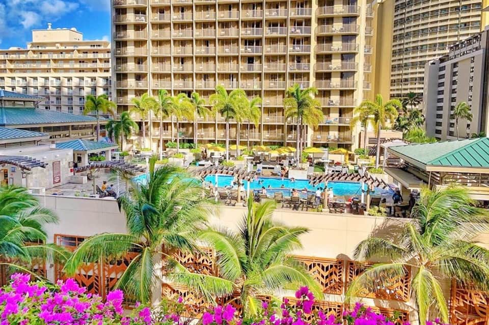 Aerial view of beachfront resort with large pool, palm trees, and high-rise hotel buildings under clear blue sky