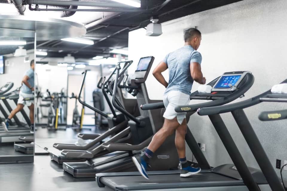 Man running on treadmill in modern gym facility with multiple cardio equipment and bright lighting