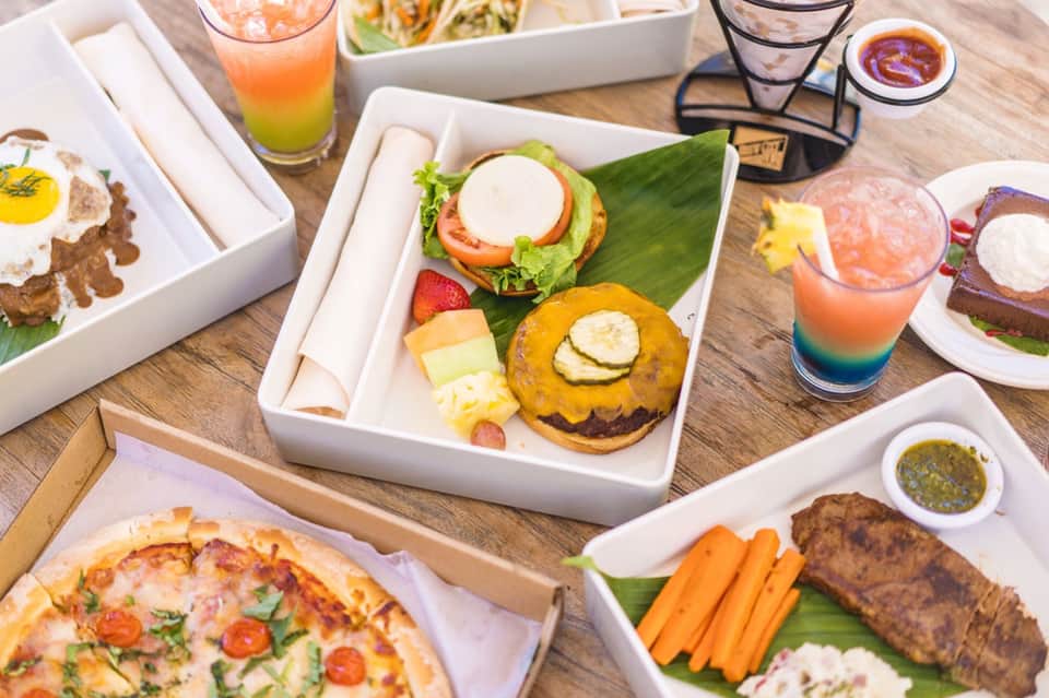 Colorful tropical dishes and beverages arranged on wooden table with pizza and grilled items