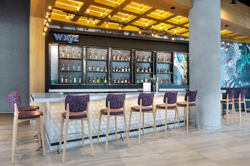 Hotel bar with illuminated wooden ceiling, purple upholstered chairs, liquor display, and contemporary design