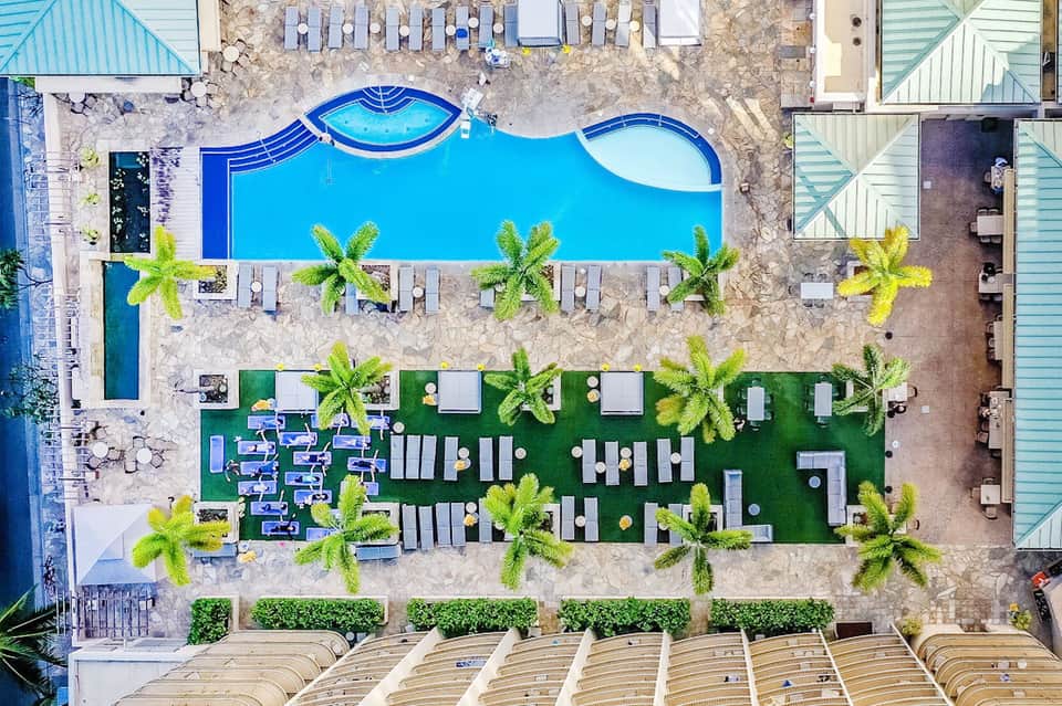 Aerial view of resort pool area with palm trees, loungers, and manicured green spaces