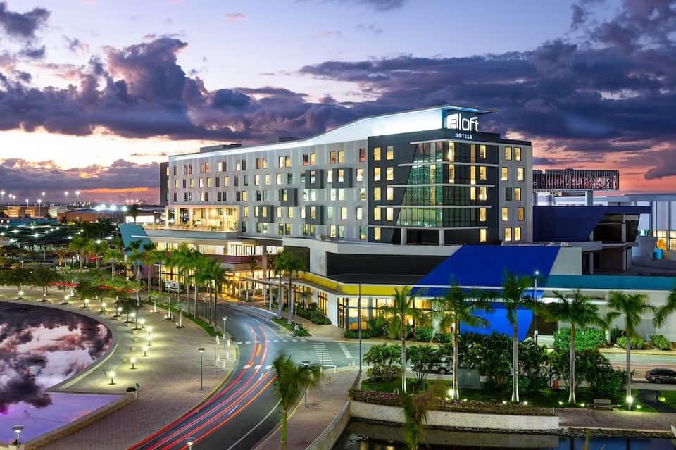Aloft hotel exterior at dusk with illuminated building, palm trees, waterfront, and modern architecture