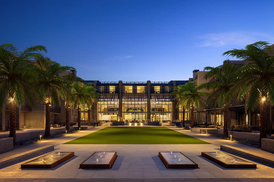 Modern hotel courtyard at dusk with illuminated brick building, centered green lawn, palm trees, and fire pits