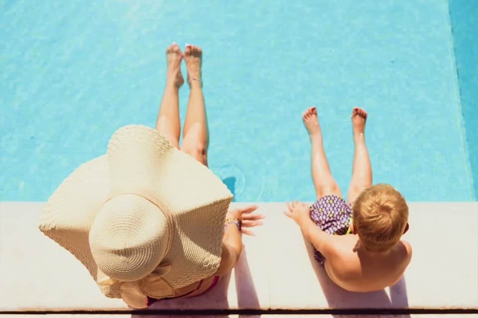 Two people dangling legs over bright blue pool edge wearing sun hats