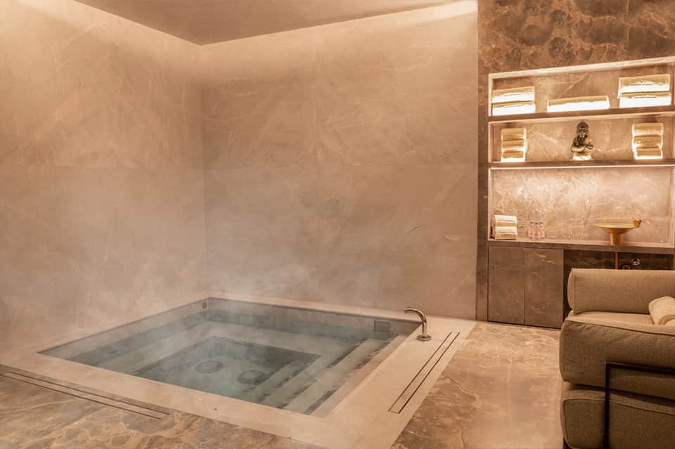 Spa treatment room with sunken soaking tub, stone walls, shelving with amenities, and ambient lighting