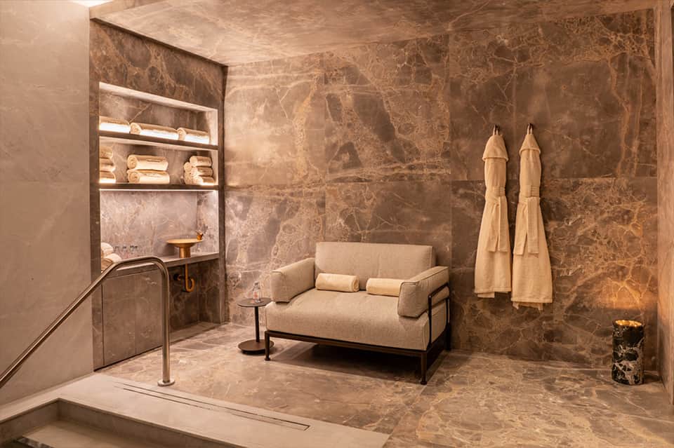 Luxury spa relaxation room with marble walls, beige lounge seating, and hanging robes with subtle lighting