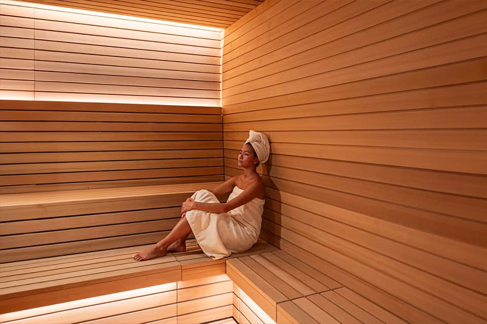 Woman relaxing in modern wooden sauna with horizontal slat walls and warm lighting