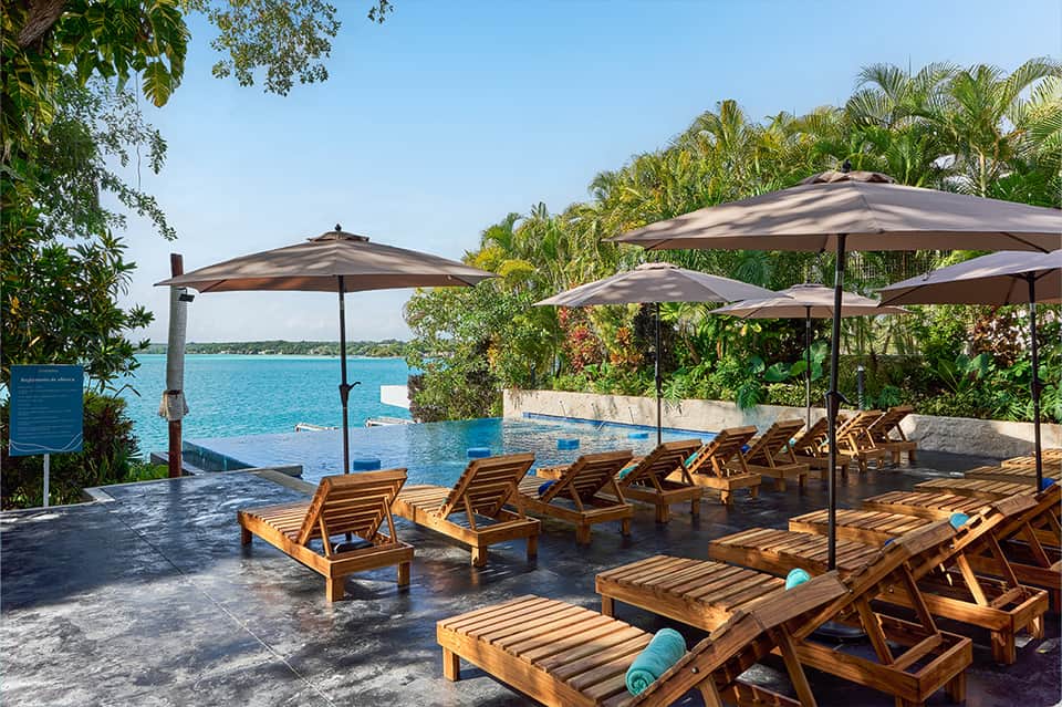 Tropical infinity pool overlooking ocean with wooden loungers, umbrellas, and palm trees