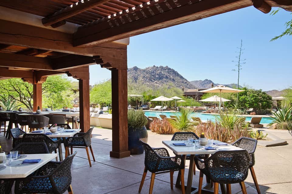 Upscale poolside dining area with wooden pergola, modern black chairs, mountain views, and umbrellas
