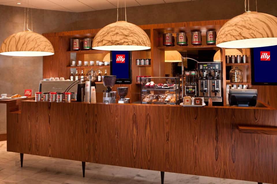 Modern coffee shop counter with wooden cabinetry, hanging dome lights, and Illy coffee branding
