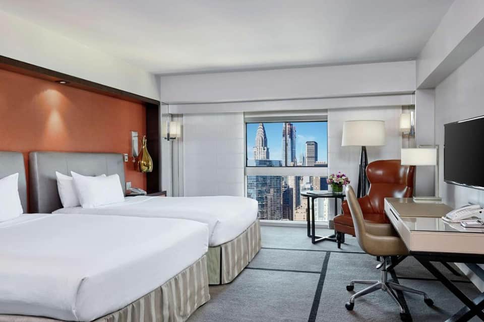 Upscale hotel room with twin beds, orange accent wall, city skyline view, and modern furnishings