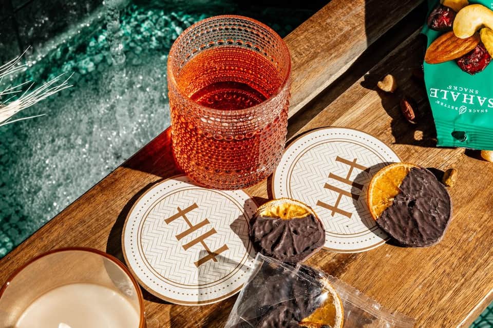 Poolside flatlay with decorative coasters, chocolate treats, nuts, coral cocktail glass, and turquoise water