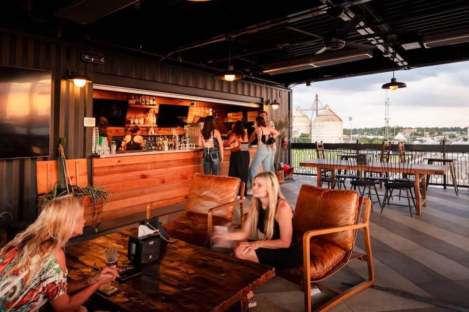 Rooftop bar at sunset with wooden seating, bar counter, and cityscape views with grain silos