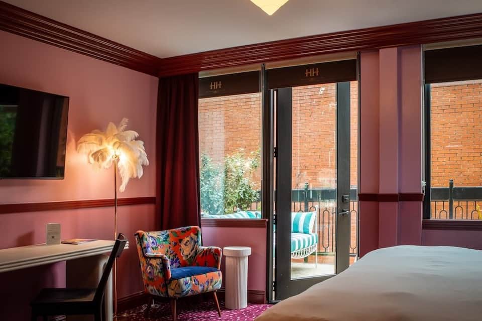 Hotel bedroom with pink walls, floral chair, palm lamp, flat-screen TV, and balcony access with brick exterior view