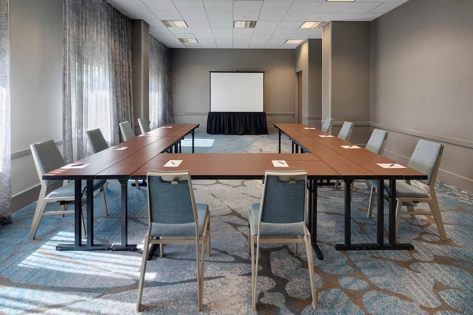 Conference room with long wooden table, chairs, projection screen, and natural light from windows