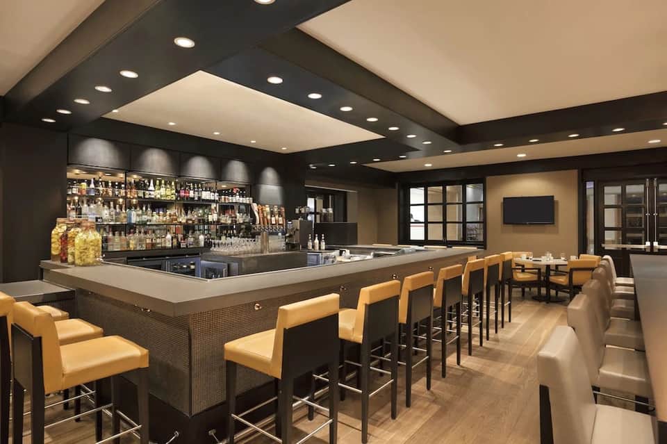 Modern bar lounge with dark wood, yellow leather stools, bottles, recessed ceiling lights, and seating area