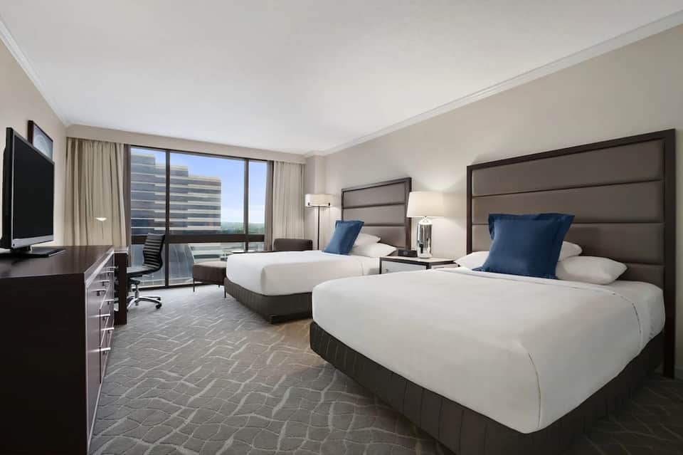 Modern hotel room with two queen beds, taupe headboards, white bedding, blue accent pillows, and city skyline views through floor-to-ceiling windows