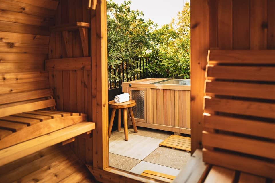 Wooden sauna entrance with hot tub visible on private outdoor deck surrounded by greenery