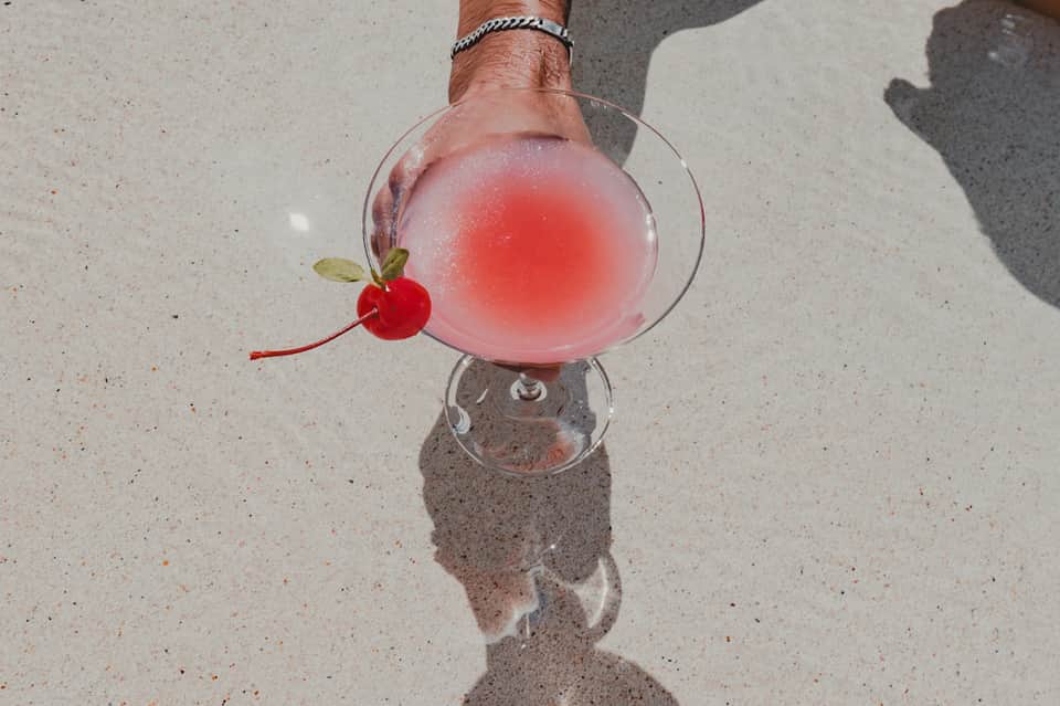Hand holding pink frozen cocktail with cherry garnish on sandy beach in bright sunlight