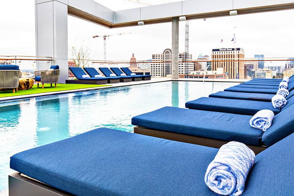 Rooftop indoor pool with city skyline views, blue loungers with white towels, and urban architecture.