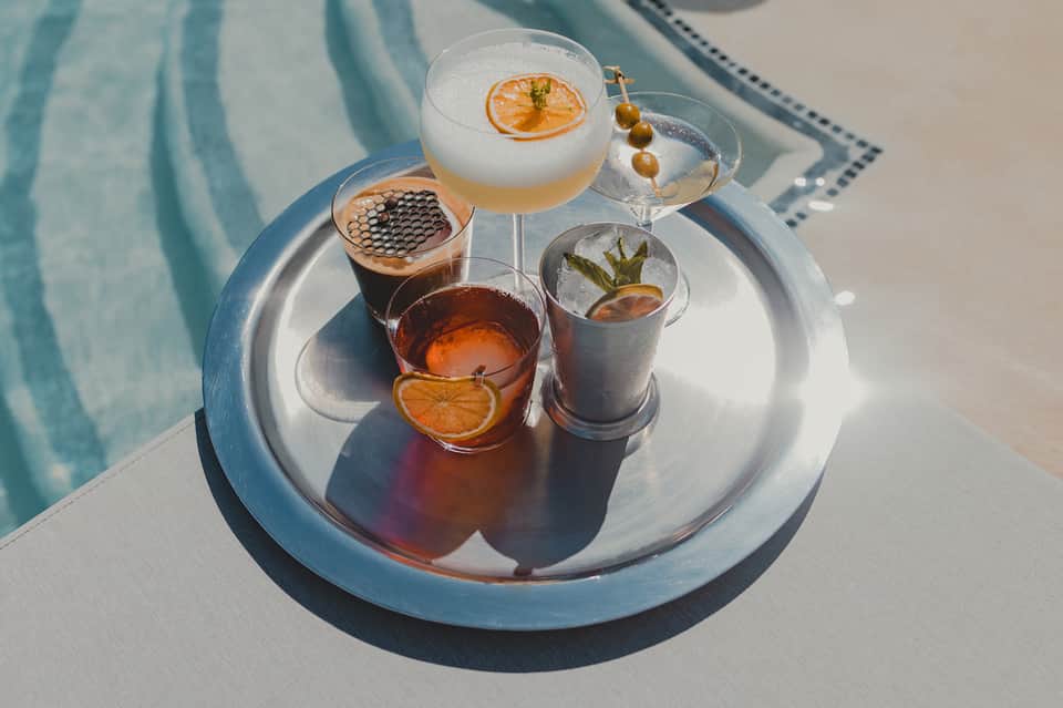 Cocktail flight on white table by pool featuring colorful drinks with citrus garnishes and blue water backdrop