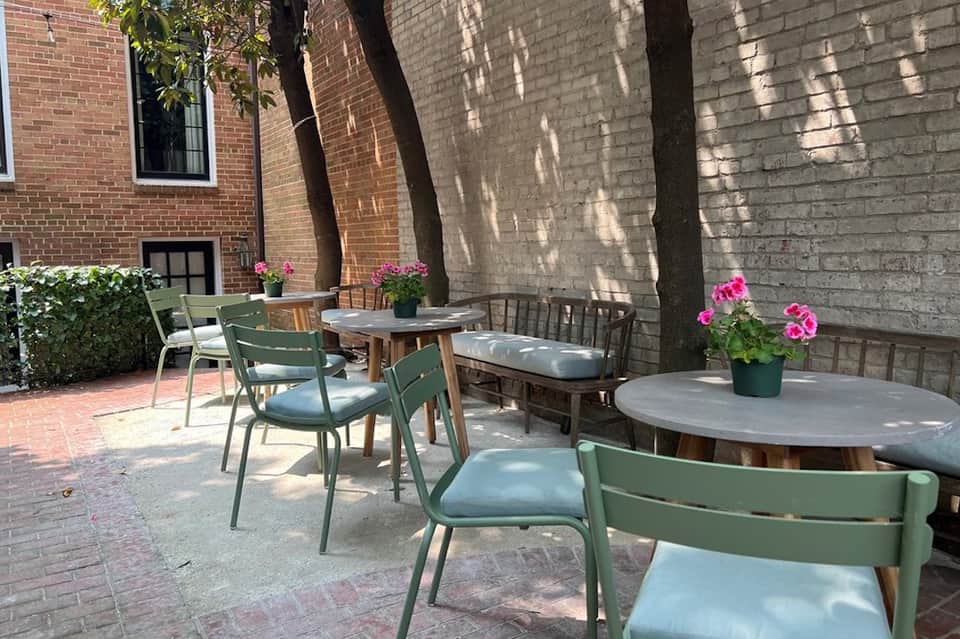 Outdoor courtyard patio with green metal chairs, round tables, and pink flowers under shade trees