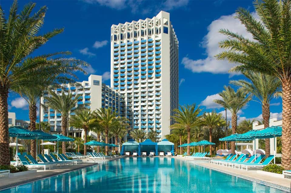 Large resort complex with turquoise pools, palm trees, lounge chairs, and high-rise hotel