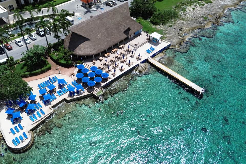 Aerial view of beachfront resort with blue umbrellas, thatched-roof structure, and pristine turquoise water
