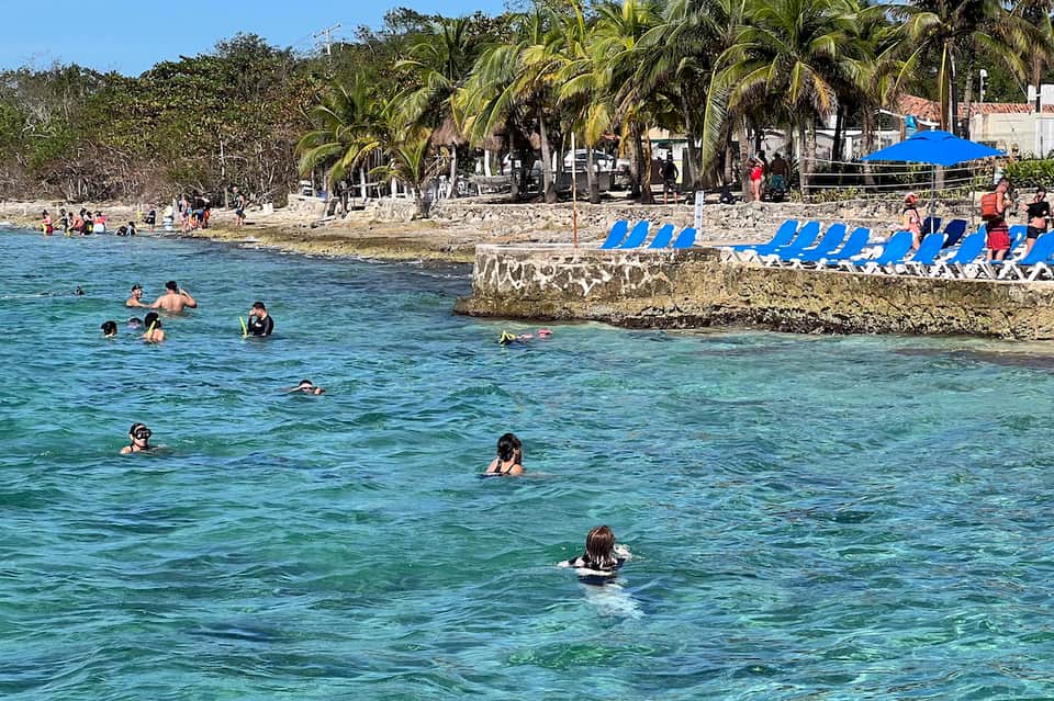 Shallow tropical beach cove with clear turquoise water, palm trees, and blue lounge chairs on shore