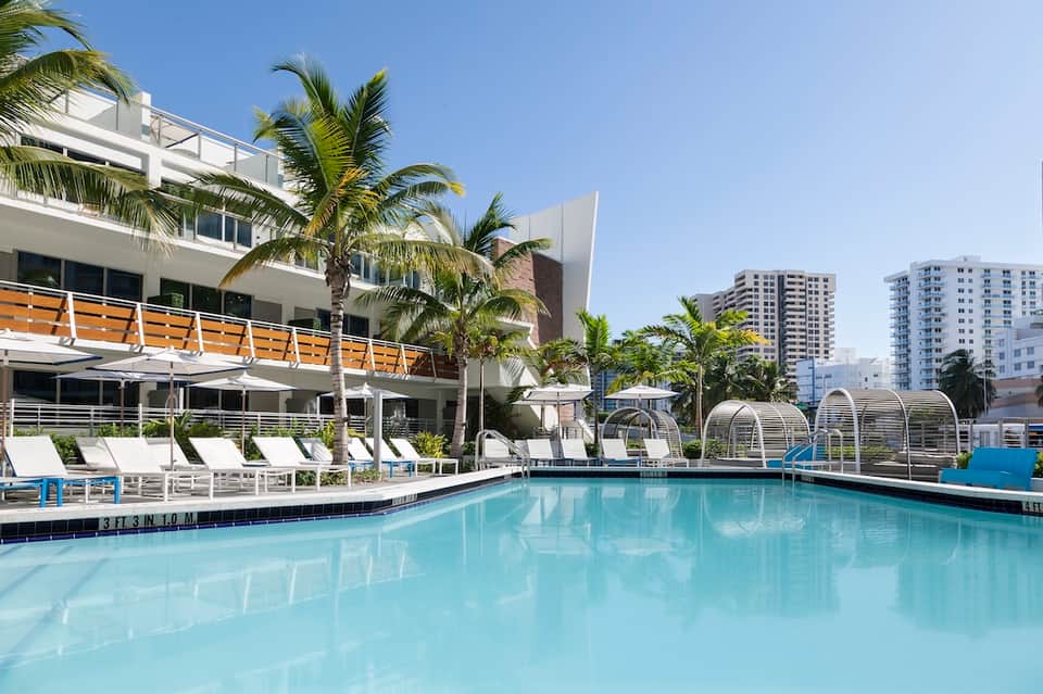 Modern waterfront pool with white loungers, palm trees, and high-rise apartment buildings