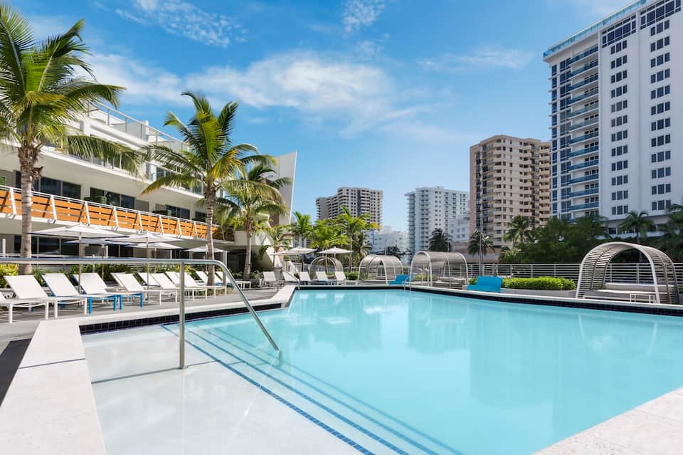 Resort pool deck with palm trees, white loungers, parasols, and multi-story residential buildings in background