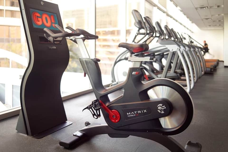Row of stationary exercise bikes with digital screens in bright fitness studio with city views