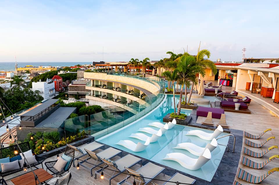 Rooftop infinity pool at sunset with ocean views, lounge chairs, palm trees, and coastal resort architecture